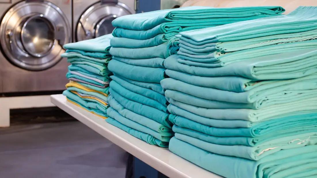 Stacks of folded teal towels on a table in a laundromat with washers in the background on the left side of the image, creating an organized laundry setup.