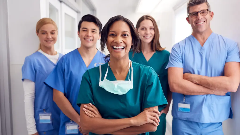Smiling healthcare team in scrubs standing in a hospital corridor, with the central nurse wearing a mask around her neck.