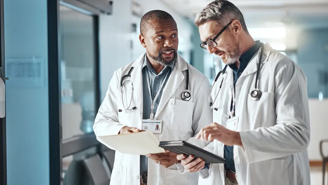 Two doctors in white coats review a folder and tablet while discussing patient information in a hospital hallway.