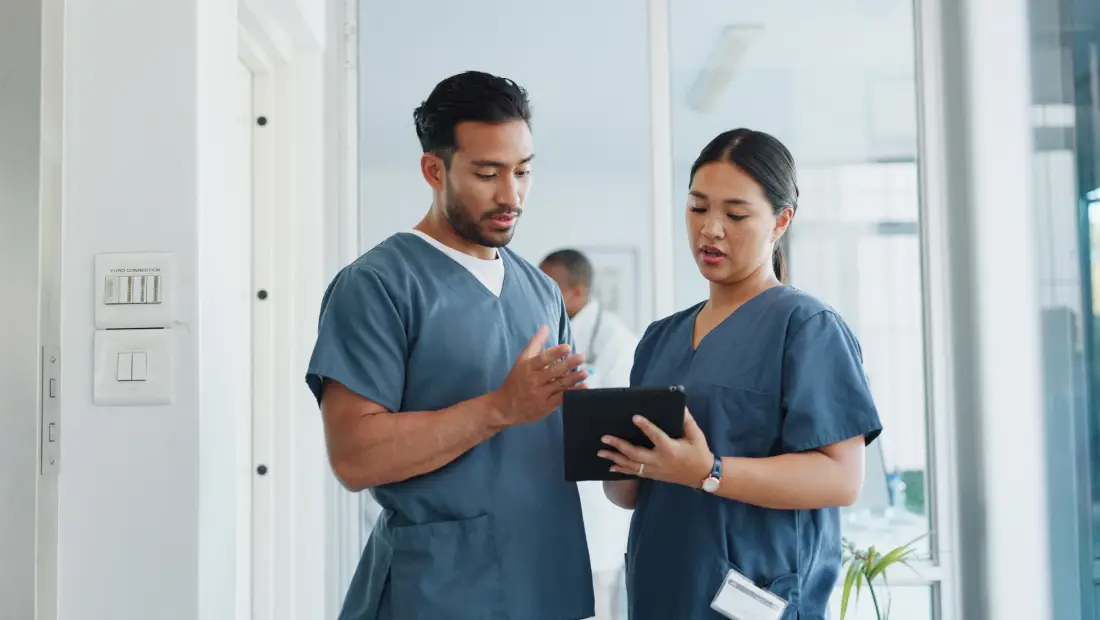 Two medical professionals in blue scrubs review information on a tablet together in a bright hospital hallway.