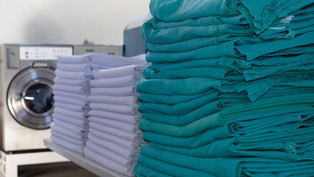 Stacks of folded teal scrubs on a table beside a stack of white scrubs, with a washing machine in the background