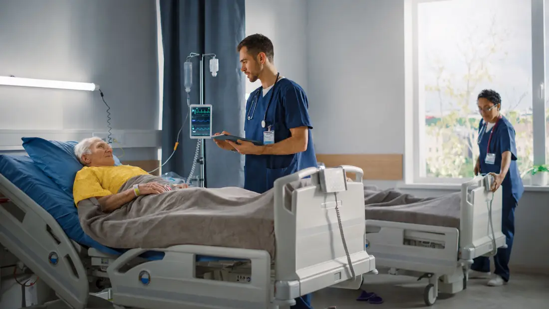 Elderly patient in a hospital bed talking to a nurse holding a tablet, with an IV drip and monitor nearby.